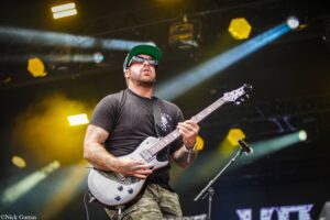 Male guitarist performing on a dim stage, wearing a green cap and sunglasses, playing a gray electric guitar amid yellow stage lights.