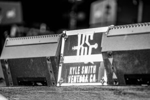 Close-up of a road case on a stage with a label reading Kyle Smith, Venice, CA (black-and-white).