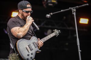 Male guitarist performing on stage, wearing a backwards cap and sunglasses, singing into a wireless mic while playing a gray electric guitar with tattoos visible on his arms.