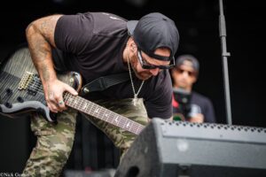 Guitarist in a black cap and sunglasses leans over a camo-patterned guitar on stage during a live performance, tattoos visible on the forearm.