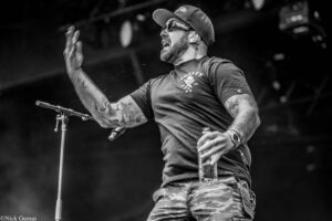 Male rock performer on stage, wearing cap, sunglasses, and tattoos, raising his hand while singing into a microphone stand.