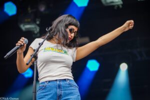 Female singer on stage with sunglasses, white Jamaica shirt, microphone in hand, arm raised high in performance.