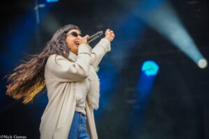 Female singer on stage, mid-performance, holding a microphone and singing with long hair and sunglasses.