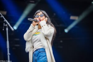 Female singer on stage, holding a microphone and singing, wearing sunglasses and a cream cardigan with blue jeans.