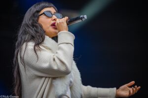 Woman singer on stage, wearing sunglasses and a cream knit sweater, singing into a handheld microphone with a dark background behind her.