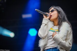 Female singer wearing sunglasses and a fluffy beige jacket, singing into a handheld microphone on stage with blue lighting behind her.
