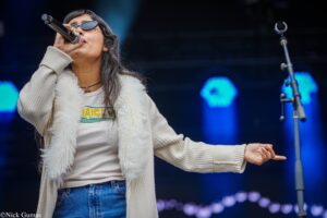 Female singer wearing sunglasses, beige cardigan, and white top sings into a microphone on stage.