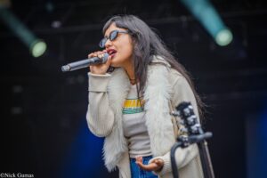Woman wearing sunglasses and a cream fur vest sings into a handheld microphone on stage, mid-performance.