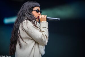 Female singer wearing round black sunglasses and a cream knit sweater sings into a handheld microphone, side profile.