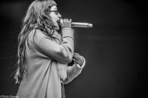 Side-profile of a female singer with long hair and glasses, singing into a handheld microphone on stage (black and white).