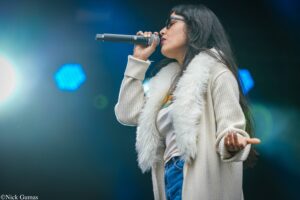 Female singer wearing glasses and a beige sweater sings into a handheld microphone on stage.