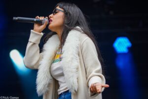 Female singer performing on stage, holding a microphone to her mouth, wearing glasses, a beige knit sweater and white fluffy scarf with blue stage lights in the background.