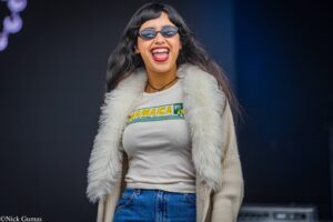 Young woman smiling broadly, wearing round sunglasses, a white fur coat, and a Jamaica graphic T-shirt.