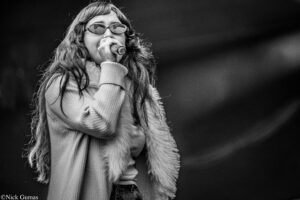 Female singer wearing sunglasses performs into a microphone on stage, with long hair and a fur vest in black and white.