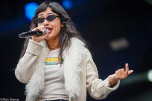 Female singer wearing sunglasses sings into a microphone on stage, wearing a cream fur vest and light sweater.