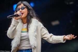A female singer wearing sunglasses and a white fur vest sings into a microphone with arms extended on stage.