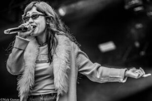 Female singer wearing sunglasses and a fur vest, singing into a handheld microphone on stage with arms outstretched in black and white. (informative)