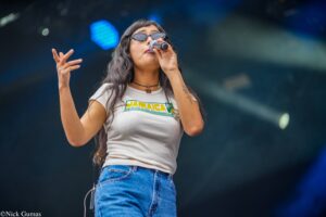 Female singer on stage, wearing sunglasses and a Jamaica shirt, holding a microphone and gesturing with her left hand while performing.
