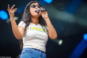 Female singer wearing sunglasses and a Jamaica-themed white shirt, singing into a microphone on stage.