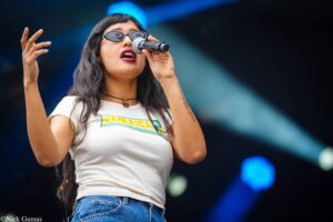 Female singer wearing sunglasses and a white Jamaica shirt, holding a microphone and performing on stage with blue light in the background.