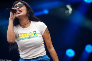 Female singer wearing sunglasses and a white Jamaica graphic shirt, singing into a handheld microphone on a dark stage with blue lighting.