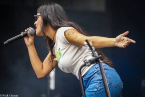 Female singer on stage, holding a microphone and extending her arm while performing, wearing sunglasses and a white top with tattoos visible on her arm.