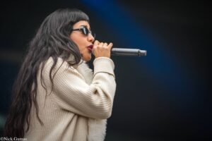 Female singer wearing sunglasses and a beige sweater, holding a microphone and singing on stage.