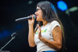 Female singer with glasses singing into a handheld microphone on stage, showing tattooed arms and a white shirt.