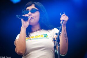 Female singer on stage, wearing sunglasses and a white shirt, singing into a handheld microphone with a blue-lit backdrop.