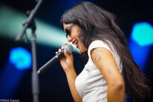 Female singer in a white top wearing sunglasses sings into a microphone on a stage with blue lights in the background.