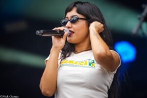 Female singer wearing round sunglasses and a white Jamaica-themed shirt, singing into a handheld microphone on stage.