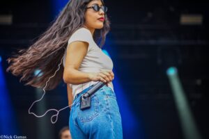 Female singer on stage wearing a white shirt and blue jeans, long dark hair flowing as she holds a microphone. She wears sunglasses and a wireless mic pack clipped to her jeans.