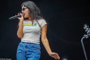 Female singer wearing sunglasses, white graphic T-shirt and jeans, holding a microphone on stage with a dark backdrop.