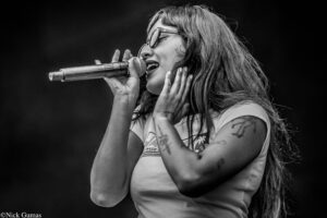 Female singer wearing glasses, singing into a microphone on a dark stage, tattoos visible on her arms photographed in black and white.