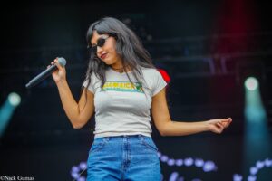 Female singer on stage wearing sunglasses, white Jamaica t-shirt and jeans, holding a microphone while performing.