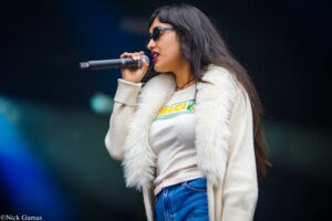 Female singer performing on stage, wearing sunglasses and a cream fur-trimmed coat, holding a microphone close to her mouth.