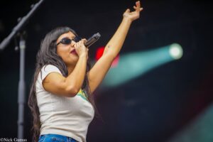 Female singer on stage, wearing sunglasses, holding a microphone and raising one arm in the air during a performance.