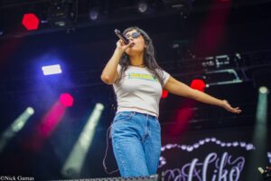Female singer on stage, microphone in hand, wearing sunglasses and a white Jamaica-themed shirt with jeans, performing under colorful stage lights.