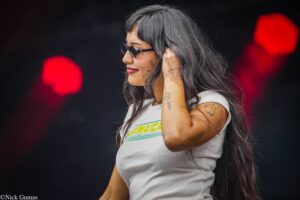 Side profile of a woman with long dark hair and tattoos, wearing round sunglasses and a white shirt, touching her hair on a stage with red lights behind her.
