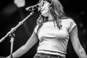 Female singer on stage, mid-performance, singing into a microphone with a wireless-looking mic stand, wearing a white T-shirt and jeans, in black-and-white.