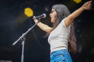 Female singer on stage singing into a microphone with arms raised, wearing a white top and blue jeans.