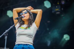 Female singer on stage, wearing sunglasses and a white t-shirt, with arms raised behind her head as she sings into a microphone.