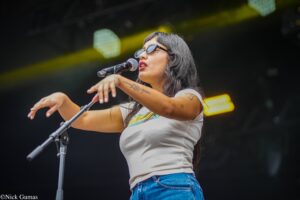 Female singer on stage wearing sunglasses, mid-performance into a handheld microphone with one arm extended.