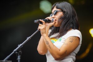 Female singer wearing sunglasses, gripping a microphone and performing on stage, with tattoos visible on her arms.