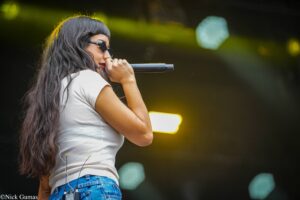Female singer on stage wearing sunglasses and a white shirt, holding a microphone toward her mouth as she performs.