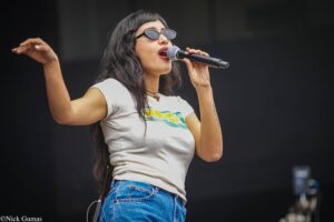 Female singer wearing sunglasses, singing into a microphone on stage with her left arm raised slightly.