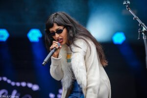 Female singer wearing sunglasses and a cream coat, singing into a microphone on a dim stage with blue lights behind her.