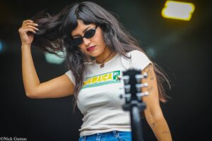 Woman with long dark hair and sunglasses adjusts her hair on stage, wearing a white Jamaica-themed t-shirt and a choker necklace, with a microphone in the foreground and stage lights in the background.