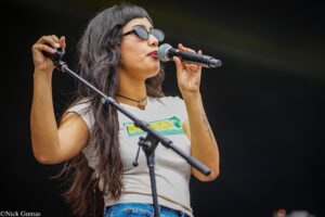 Female singer with long dark hair and sunglasses singing into a handheld microphone on a dark stage.