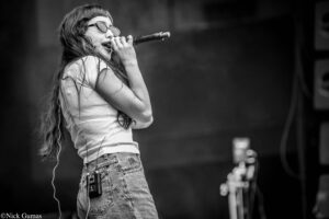 Female singer with long hair and glasses, singing into a handheld mic on stage, wearing a light top and jeans with wireless pack on her belt.
